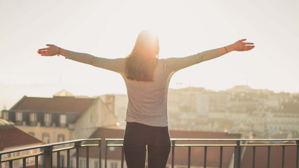Perempuan berdiri di balkon sambil merentangkan tangan menikmati sinar matahari pagi